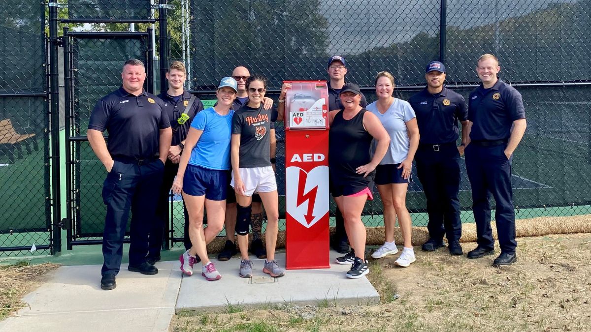 Group of firefighters and laypeople standing around red AED installation in front of tennis courts.