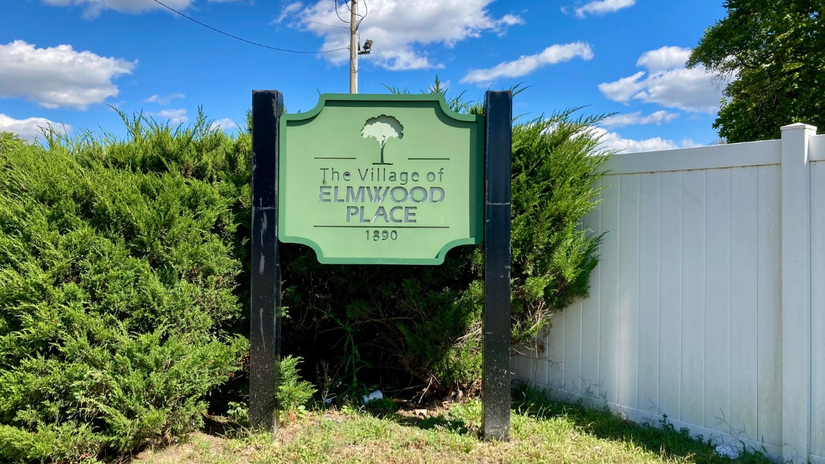 Sign saying The Village of Elmwood Place 1890 in front of green bushes and a white fence.