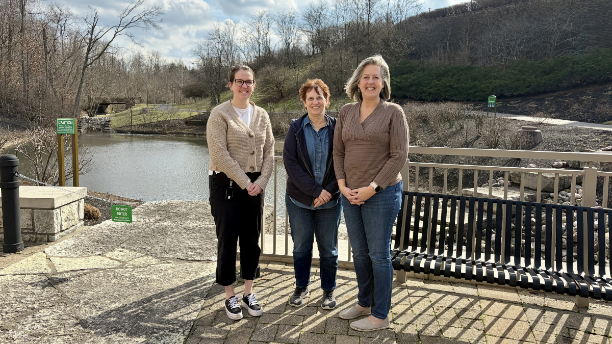 Three females posing in front of pond in a park like setting.