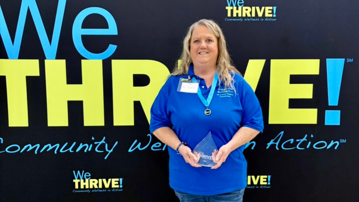 Pam Jackson holding the champion award standing behind a large WeTHRIVE! banner.