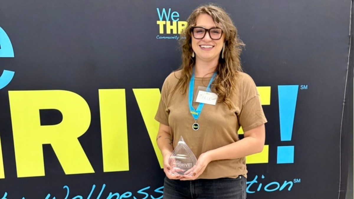 Sable Bender holds WeTHRIVE! champion award, smiling, while standing in front of a large WeTHRIVE! banner.